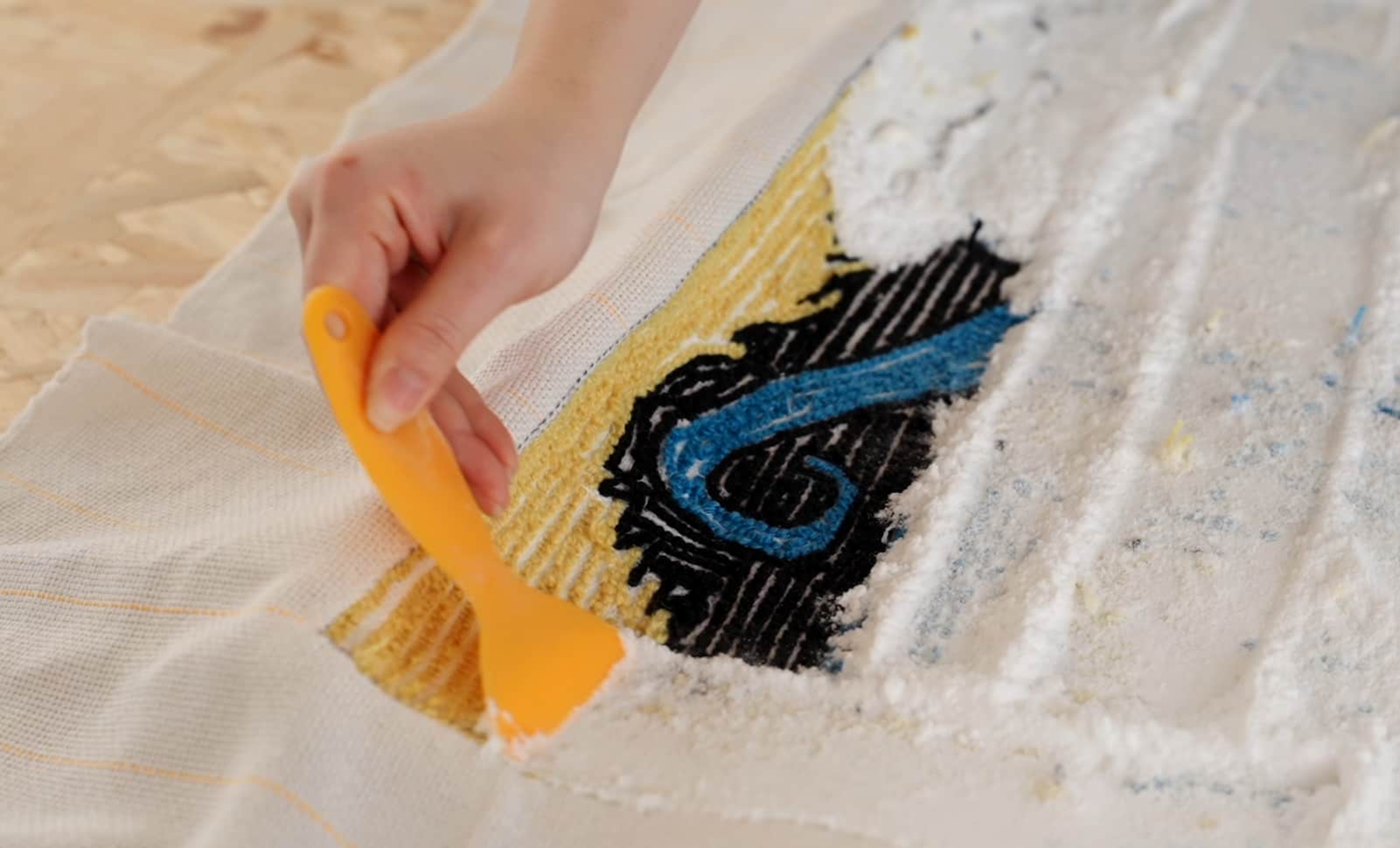 A close-up shot of a hand using a yellow plastic spatula to spread white rug adhesive evenly over the back of a colorful tufted design on canvas.