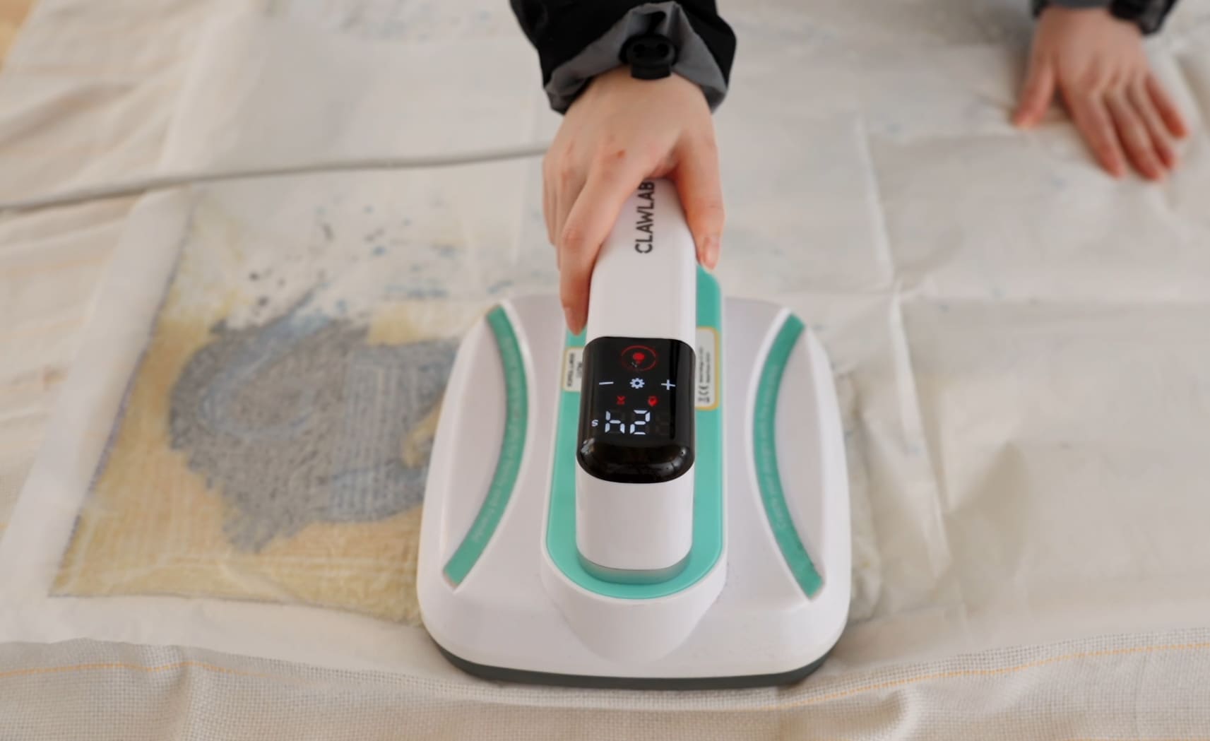 Close-up of a hand using a white and teal CLAWLAB heat press on the back of a tufted design through a protective sheet. The device's digital screen displays 24.
