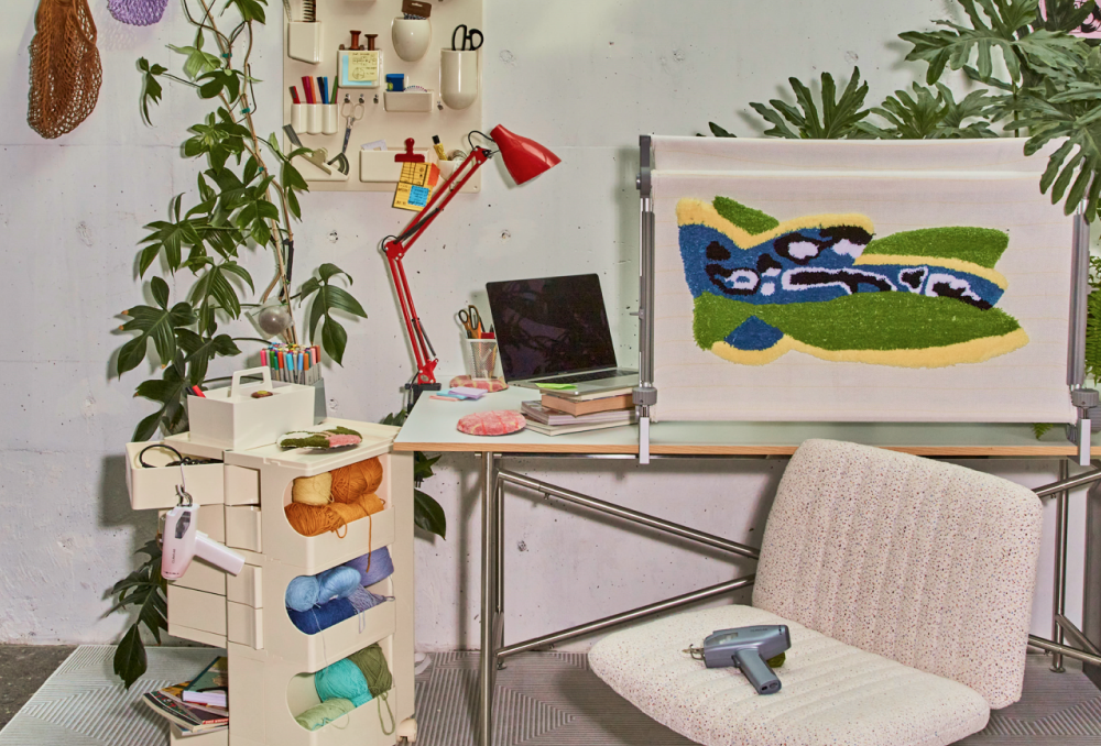 Cozy tufting workspace with a desk, yarn storage cart, a Clawlab tufting gun, tufted artwork on a frame, and a cushioned chair.