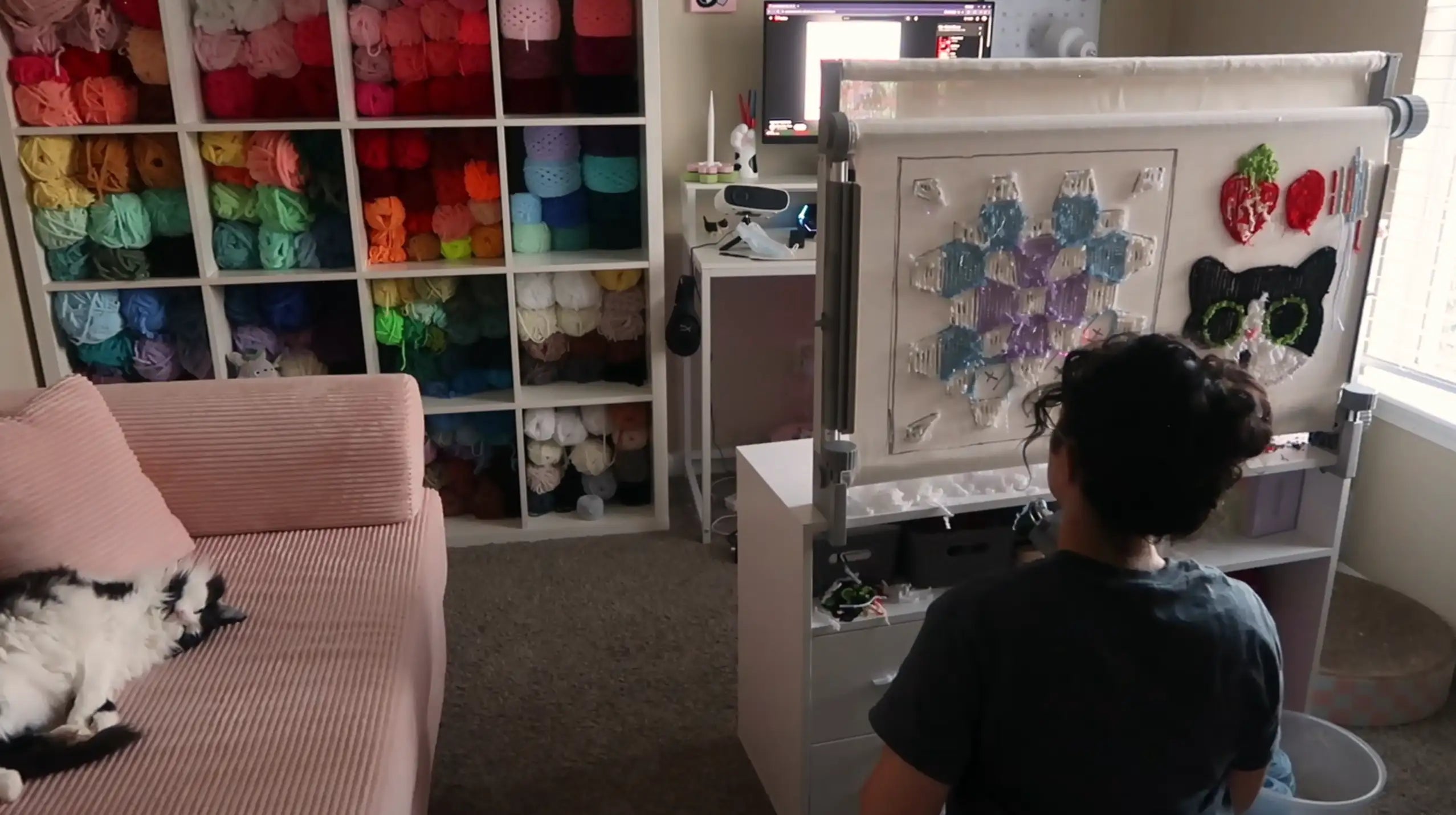 Person using a tufting gun at a frame in a craft room with colorful yarn shelves and a cat on a sofa ​