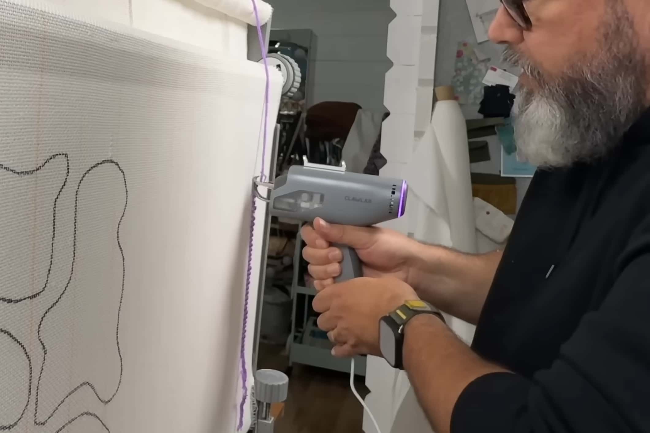A man with a beard uses a grey Clawlab tufting gun to create a purple outline on a white canvas frame, demonstrating a focused and creative DIY crafting session.