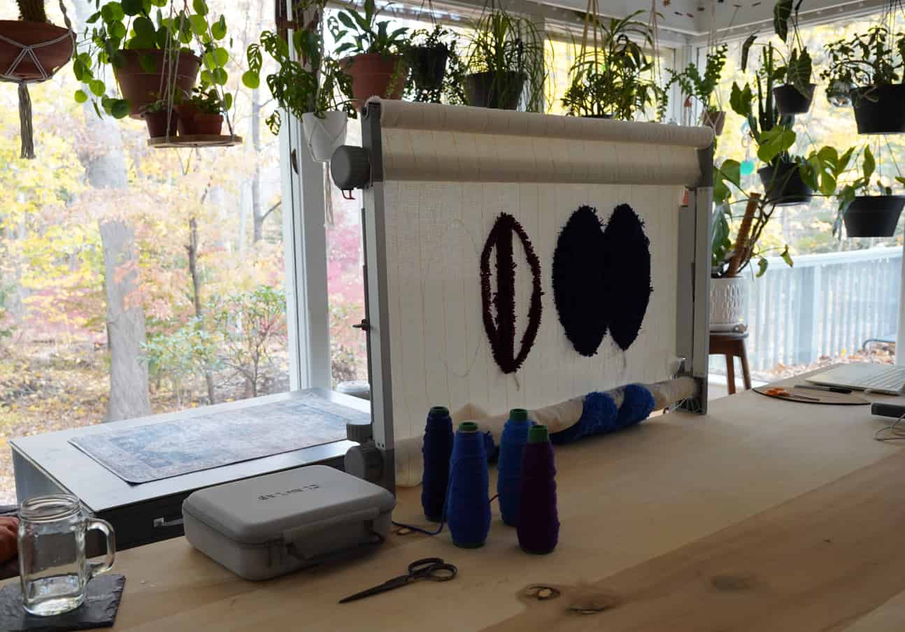 A tufting frame in a sunroom with three dark oval shapes partially tufted on the canvas, surrounded by cones of blue and purple yarn on a wooden table.