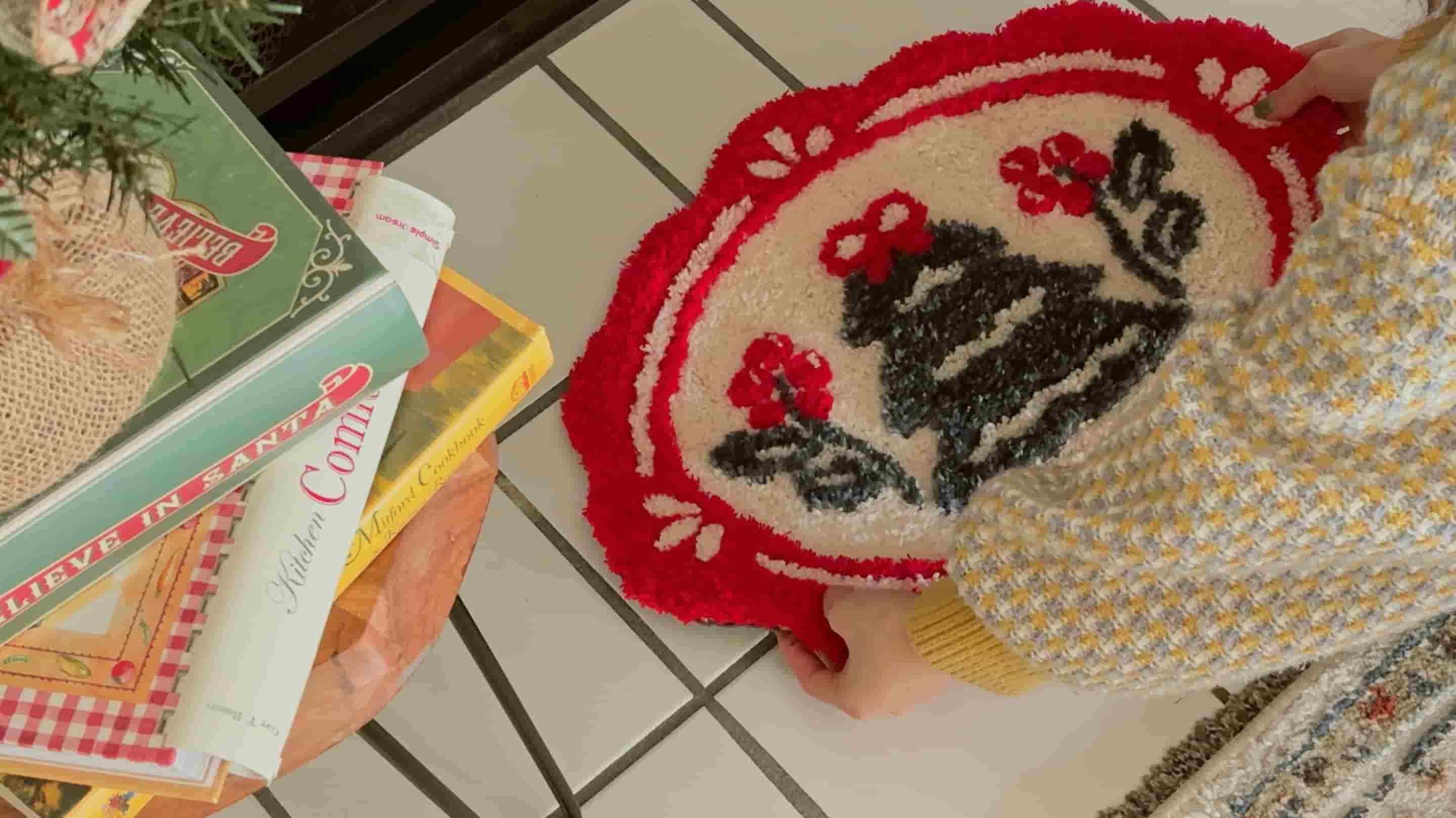 A person holds a red-and-white round tufted rug with a black cat and red flowers on a tiled floor beside a stack of holiday-themed books and cards.