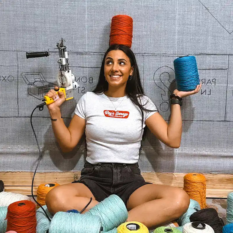 A smiling woman holding a tufting gun and colorful yarn spools in a home studio setup.