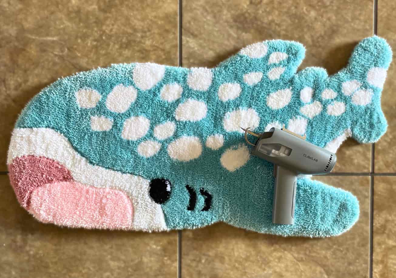 A finished custom whale shark shaped tufted rug, combining turquoise, pink, white, and black colors, with a Clawlab tufting gun resting on the floor beside it.