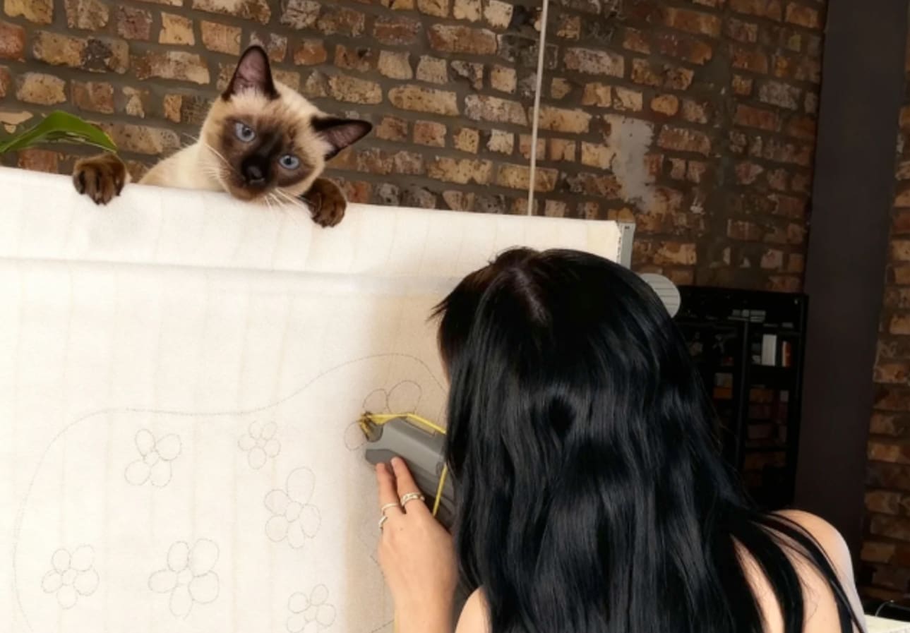 Siamese cat perched on a tufting frame watching a woman use a tufting gun.