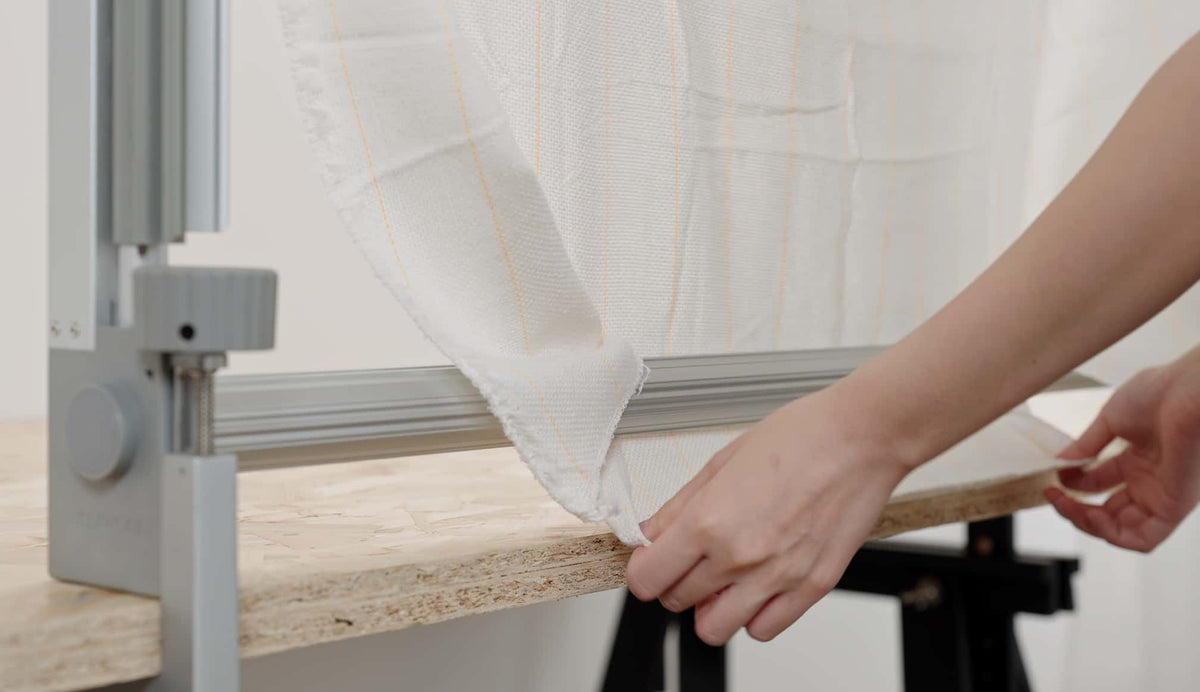 Hands securing white tufting fabric with yellow stripes onto Clawlab tufting frame for precise results.